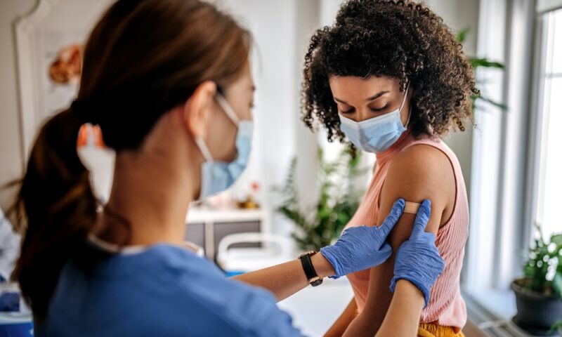 A nurse putting adhesive bandage on young woman patient’s arm after vaccination at a clinic.