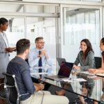 Smiling business team discussing work in meeting using laptop and documents in conference room. Successful business woman and businessmen discussing marketing strategy and ideas at meeting room. Corporate business man and woman with manager and secretary working together.