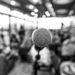 Black and white photo of a microphone infront of defocused people indoors