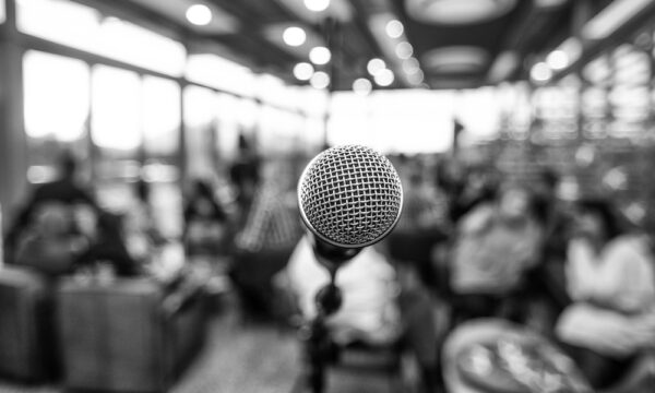 Black and white photo of a microphone infront of defocused people indoors