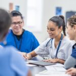 A small group of medical professionals gather around a table as they meet together to discuss patient cases. They are each dressed professionally and have files scattered between them for reference.