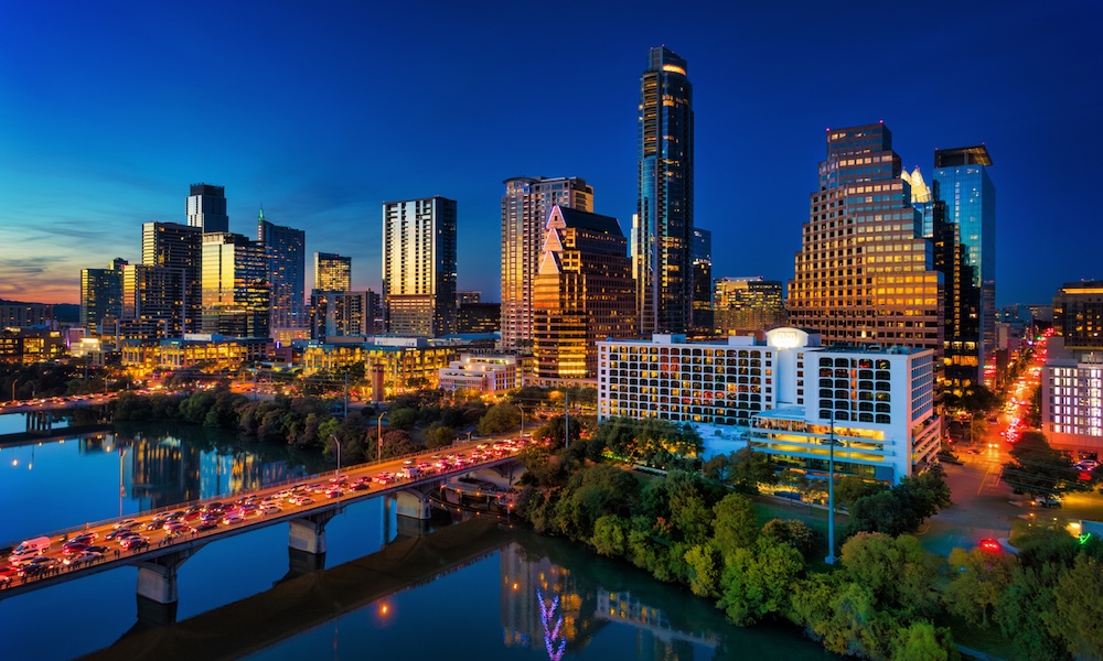 Downtown Austin skyline, elevated view, at dusk with the sunset reflections on the building facade, and the Lady Bird Lake / Colorado River with heavy traffic on the bridges in the foreground.