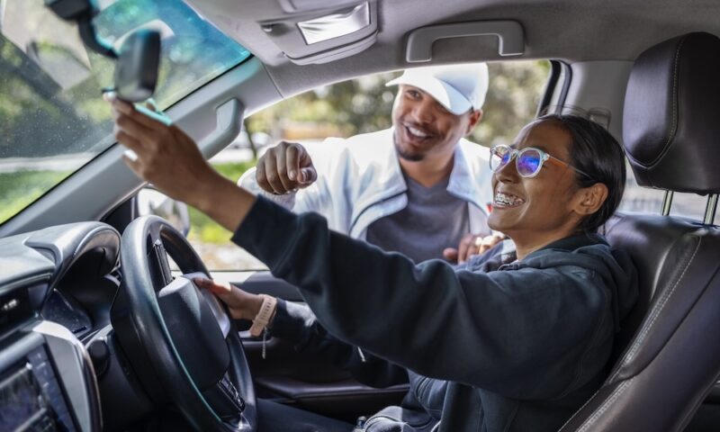 Cheerful female student adjusting car rear view mirror during driving lesson with male instructor