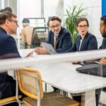 Multiracial governing board meeting at conference table, shot through window. Businessmen and businesswomen planning company strategy, medium shot