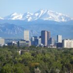 Snow covered Longs Peak, part of the Rocky Mountains stands tall in the background with green trees and the Downtown Denver skyscrapers as well as hotels, office buildings and apartment buildings filling the skyline.