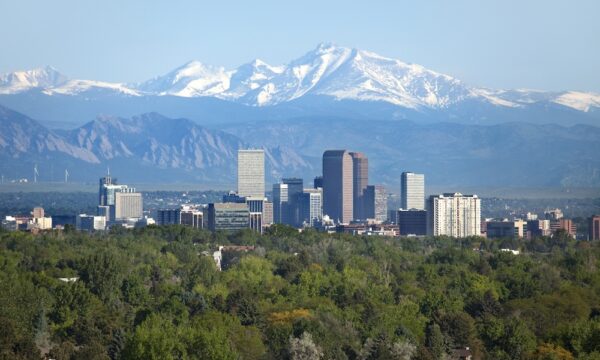 Snow covered Longs Peak, part of the Rocky Mountains stands tall in the background with green trees and the Downtown Denver skyscrapers as well as hotels, office buildings and apartment buildings filling the skyline.