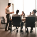 Business Presentation, Blurred Background. Businessman Giving Speech During Seminar With Coworkers In Office, Standing At Desk In Boardroom, Diverse People Sitting At Table And Listening To Speaker